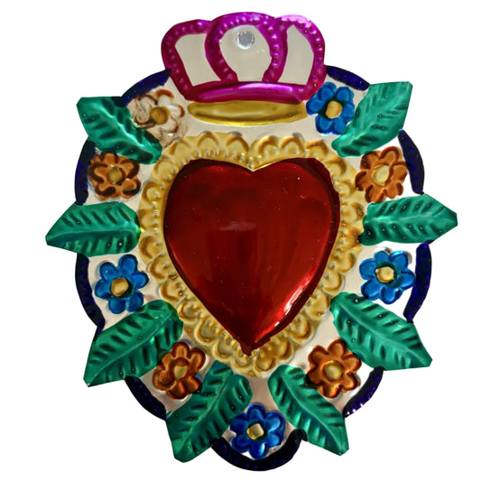 A colorful Mexican-style metal heart ornament featuring a red heart in the center, surrounded by a gold scalloped border, green leaves, blue and orange flowers, and a pink crown at the top, displayed on a plain white background.